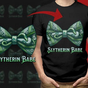 Może przedstawiać: Czarna koszulka z zielonym wzorem muszki z motywem węża i napisem "Slytherin Babe" w kolorze zielonym. Wzór muszki powtarza się w tle.