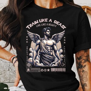 Può includere: T-shirt nero con grafica di un uomo muscoloso con ali, barba e drappeggio. Il testo "TRAIN LIKE A BEAST LIVE LIKE A BEAUTY" è sopra l'immagine. Il testo aggiuntivo include "BUSY LIFTING" e "ESTD. 2024".