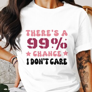 Könnte beinhalten: Weißes T-Shirt mit dem Text "THERE'S A 99% CHANCE I DON'T CARE" in Rosa, Dunkelrosa und Schwarz. Der Text ist in einer Retro-Schriftart gehalten. Das T-Shirt hat einen lässigen Stil.