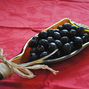 May include: A glass serving dish shaped like a bottle, filled with black olives. The dish is amber-colored with a gold rim and tied with raffia. The background is a red tablecloth.