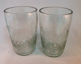 May include: Two clear glass tumblers with a floral embossed design. The glasses have a slightly tapered shape and a smooth rim. The embossed pattern covers the lower half of the glasses.