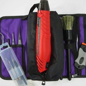 Op de afbeelding: Een zwarte en paarse gereedschapsorganizer met een rode heteluchtpistool, borstels en diverse gereedschappen. De organizer heeft meerdere vakken en een ritssluiting. De gereedschappen omvatten pincetten, schrapers en andere kleine instrumenten, ideaal voor knutselen of reparaties.