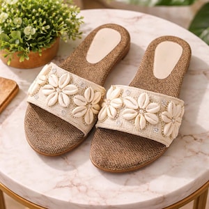 Puede incluir: Un par de sandalias beige y marrón con un diseño floral. Las sandalias tienen una base marrón tejida, una tira beige adornada con adornos blancos en forma de flor y pequeñas cuentas plateadas. Las sandalias se exhiben sobre una mesa con tapa de mármol.