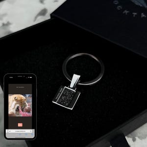 Könnte beinhalten: Ein silberner Schlüsselanhänger mit einem quadratischen Anhänger mit einem QR-Code und dem Text "JEWEL COCKTAIL". Der Schlüsselanhänger wird in einer schwarzen Box präsentiert. Ein Smartphone zeigt ein Foto einer Person, die einen Löwen berührt.