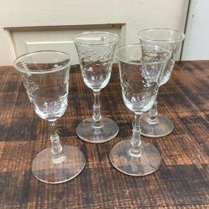 Könnte beinhalten: Vier klare Weingläser mit floralen Ätzungen. Jedes Glas hat einen runden Fuß, einen detaillierten Stiel und eine ausgestellte Schale. Die Gläser stehen auf einer dunklen Holzoberfläche und zeigen ihre filigrane Handwerkskunst.