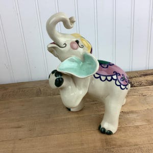 Puede incluir: Una figura de elefante de cerámica crema, con detalles coloridos. El elefante tiene una trompa levantada, orejas grandes y está decorado con detalles rosas, amarillos, verdes y azules. La figura está sobre una superficie de madera.