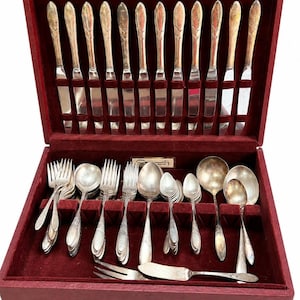 May include: A silver-toned cutlery set presented in a rectangular red velvet-lined wooden case. The collection features twelve knives with patterned handles, along with assorted forks, spoons, and a serving utensil. A small metal plaque marked Community is visible.