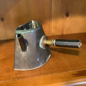 Puede incluir: Jarra antigua de cobre y latón con un pico y un asa geométrica negra. La jarra tiene un acabado patinado, que muestra signos de edad y uso. El asa está unida con un accesorio de latón.