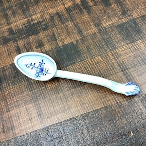 May include: A white ceramic spoon with a blue floral design in the bowl and on the handle's end. The spoon is curved and rests on a dark brown wooden surface. The spoon's bowl is oval-shaped.