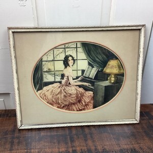 Peut inclure: Impression encadrée d'époque d'une femme jouant du piano. L'œuvre représente une femme en robe rose, assise devant un piano, avec une scène hivernale visible à travers une fenêtre. Une lampe est posée sur une petite table à côté du piano.