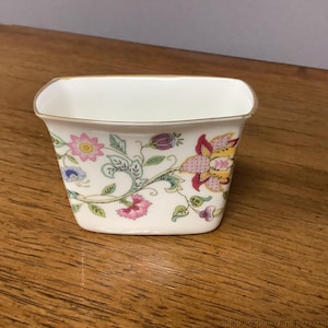 Könnte beinhalten: Eine kleine, rechteckige Porzellanschale mit floralem Design. Die weiße Schale zeigt rosa, gelbe und lila Blumen mit grünen Blättern. Der Rand ist mit Gold verziert. Die Schale steht auf einer Holzoberfläche.
