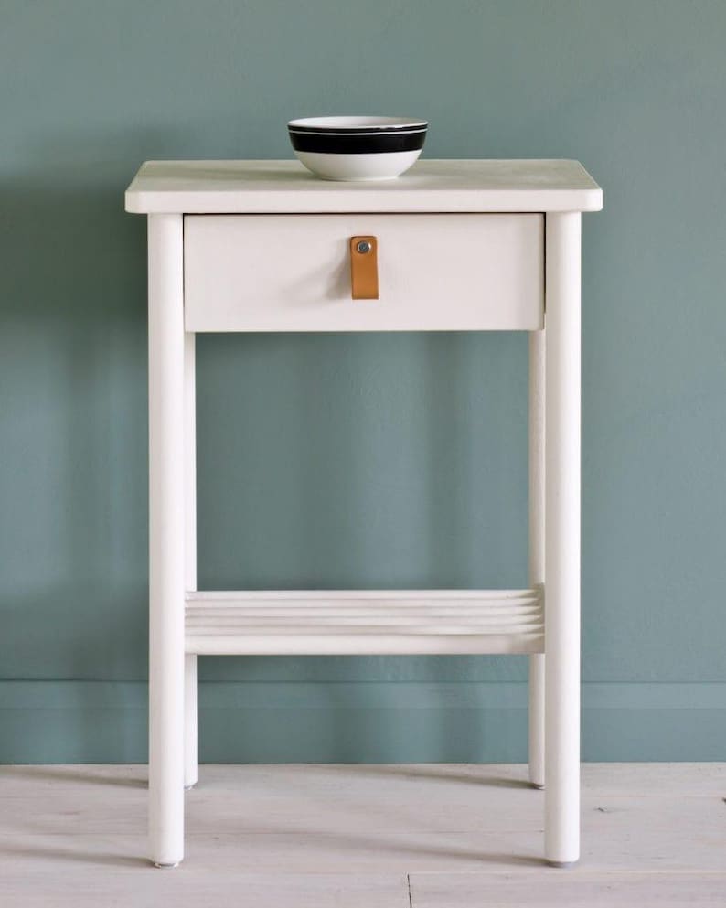 May include: A white bedside table with a single drawer and a small shelf. The drawer has a tan leather pull. A small white bowl with a black stripe is on top. The table is approximately 24 inches tall.