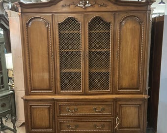 Könnte beinhalten: Ein großer brauner Holzschrank mit zwei oberen Schranktüren mit goldenen Metallgittereinsätzen, zwei massiven Holztüren an den Seiten und zwei unteren Schubladen mit verzierten Metallgriffen. Die Oberseite ist gewölbt und mit Schnitzereien versehen.