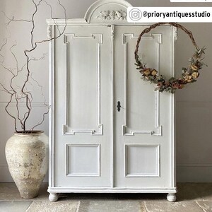 May include: A white, antique-style cabinet with decorative molding and a floral wreath hanging on the door. A large, aged ceramic vase with branches is to the left. The cabinet has a classic design and is painted in a neutral color.