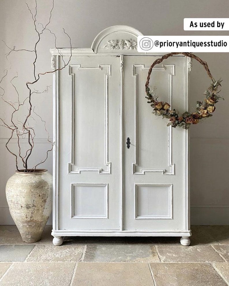 May include: A white, antique-style cabinet with decorative molding and a floral wreath hanging on the door. A large, aged ceramic vase with branches is to the left. The cabinet has a classic design and is painted in a neutral color.