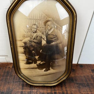 Puede incluir: Fotografía antigua en tonos sepia enmarcada en un marco dorado de forma inusual. La imagen muestra a dos personas sentadas en unos escalones. Los tonos sepia sugieren una pieza antigua, capturando un momento del pasado.