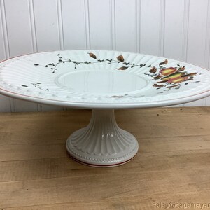 May include: White ceramic cake stand with a fluted edge and a floral design. The stand has a pedestal base with a red trim.
