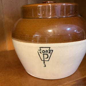 May include: A vintage stoneware crock with a brown glazed top and a white base. The crock features a black emblem with the word "YORK" and the letter "P", along with the number "3".