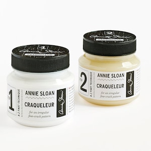Könnte beinhalten: Zwei Gläser Annie Sloan Craqueleure, eine 2-teilige Technik zur Erstellung eines unregelmäßigen Feinsprungmusters. Ein Glas ist weiß, mit der Aufschrift "1", das andere ist beige, mit der Aufschrift "2". Beide haben schwarze Deckel.