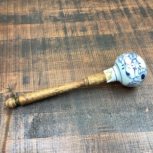 May include: A vintage-style, hand-held object with a wooden handle and a ceramic head. The ceramic head is white with blue floral patterns. A green string is attached to the wooden handle. The object is placed on a wooden surface.