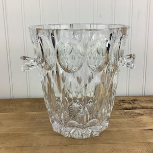 Pode incluir: Um balde de gelo de vidro transparente e facetado com duas alças. O balde tem uma parte superior larga e aberta e um padrão decorativo de formas ovais e cortes de diamante. A base tem um design de estrela. Projetado para conter gelo para resfriar bebidas.