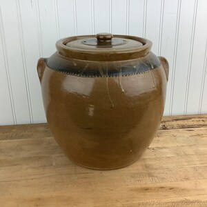 May include: A large, glazed stoneware crock with a lid. The crock is a warm brown color with a darker band around the top and two handles. The lid has a small knob. The crock is sitting on a wooden surface.