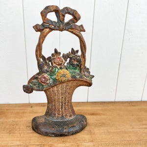 May include: A cast iron doorstop shaped like a basket of flowers with a prominent bow handle. The piece features a textured woven basket base, green leaves, and multi-colored floral accents in red, yellow, and dark tones with a weathered finish.