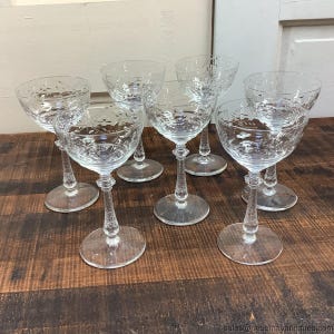 Könnte beinhalten: Eine Sammlung von acht transparenten Champagnergläsern. Jedes Glas hat eine runde Schale mit geätzten floralen Mustern, einen strukturierten Stiel und einen kreisförmigen Fuß. Die Gläser stehen auf einer dunklen Holzoberfläche und zeigen ihr elegantes Design.