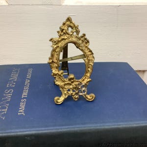 Puede incluir: Un pequeño marco de fotos dorado y ornamentado con una abertura ovalada. El marco tiene un diseño decorativo de volutas y se apoya en un pequeño caballete. El marco está sobre un libro azul. El marco es probablemente vintage o antiguo.