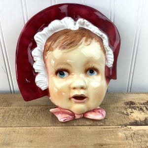 May include: A ceramic wall plaque depicting a child's face wearing a red bonnet and a pink bow. The child has blue eyes and a fair complexion.