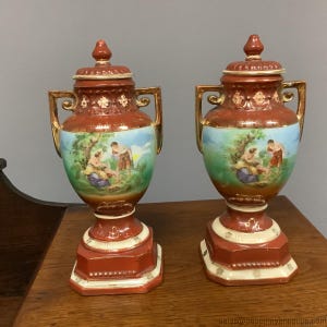 Może przedstawiać: Para antycznych ceramicznych urn z pokrywkami. Urny są głównie czerwonobrązowe ze złotymi akcentami i uchwytami. Każda urna przedstawia malowaną scenę postaci w krajobrazie. Podstawy to połączenie kolorów czerwonego i kremowego.