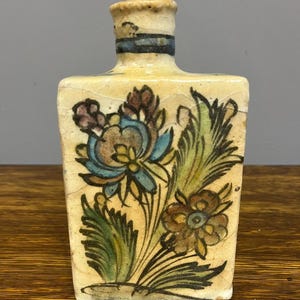 May include: A small, rectangular ceramic bottle with a cream-colored base and floral design. The flowers are in shades of blue, brown, and green, with black outlines. The bottle's neck has a blue band. The piece is likely vintage or antique.