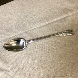May include: A silver spoon with an ornate handle. The spoon is lying on a beige fabric surface.