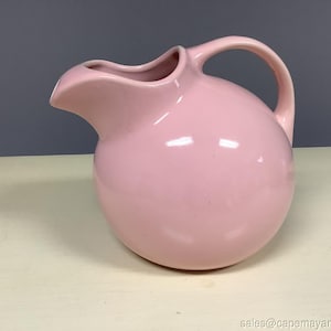 Puede incluir: Una jarra de cerámica rosa claro vintage con un cuerpo redondeado y un asa curvada. La jarra tiene un pico para verter y probablemente se utiliza para servir bebidas. La jarra está sobre una superficie de color claro.