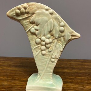 May include: A vintage ceramic vase in a fan shape, with a relief design of grapes and leaves. The vase has a light green and tan colour scheme, with a rectangular base. The design is detailed and the vase appears to be an antique.