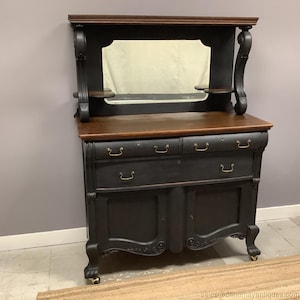 May include: An antique black buffet with a mirror, two drawers, and two doors. The buffet has a dark wood top and a decorative mirror. The hardware is brass-colored. The legs have brass-colored accents.