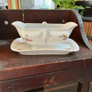 May include: A white ceramic gravy boat with an attached underplate, featuring delicate hand-painted pink and yellow floral motifs. The gravy boat has a curved handle and a wide pouring spout, while the rectangular base plate matches the floral pattern.