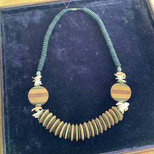 Puede incluir: Un collar artesanal con cuentas de madera oscuras y claras. El collar tiene un diseño curvo con colgantes redondos de madera grandes y detalles de conchas marinas. El collar se exhibe sobre una superficie azul oscuro.