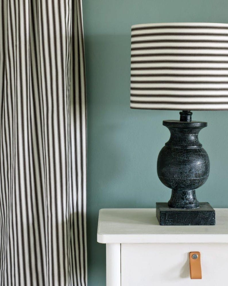 May include: A black table lamp with a striped lampshade sits on a white nightstand. The lamp has a textured, urn-shaped base. A matching striped curtain hangs to the left. The background is a muted teal color.