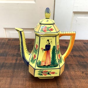 May include: A yellow ceramic teapot with a lid, spout, and handle. The teapot is decorated with a woman in traditional clothing, flowers, and green trees. The spout is blue, and the handle is orange.
