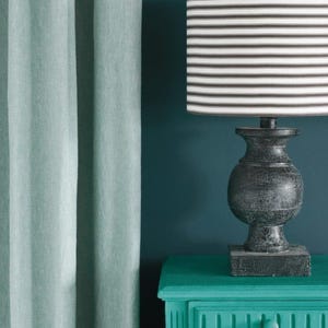 K&ouml;nnte beinhalten: Eine dekorative Lampe mit einem schwarz-wei&szlig; gestreiften Lampenschirm steht auf einem t&uuml;rkisfarbenen Nachttisch. Die Lampe hat einen dunklen, strukturierten Sockel. Weiche t&uuml;rkisfarbene Vorh&auml;nge sind links, der Hintergrund ist tief t&uuml;rkis.