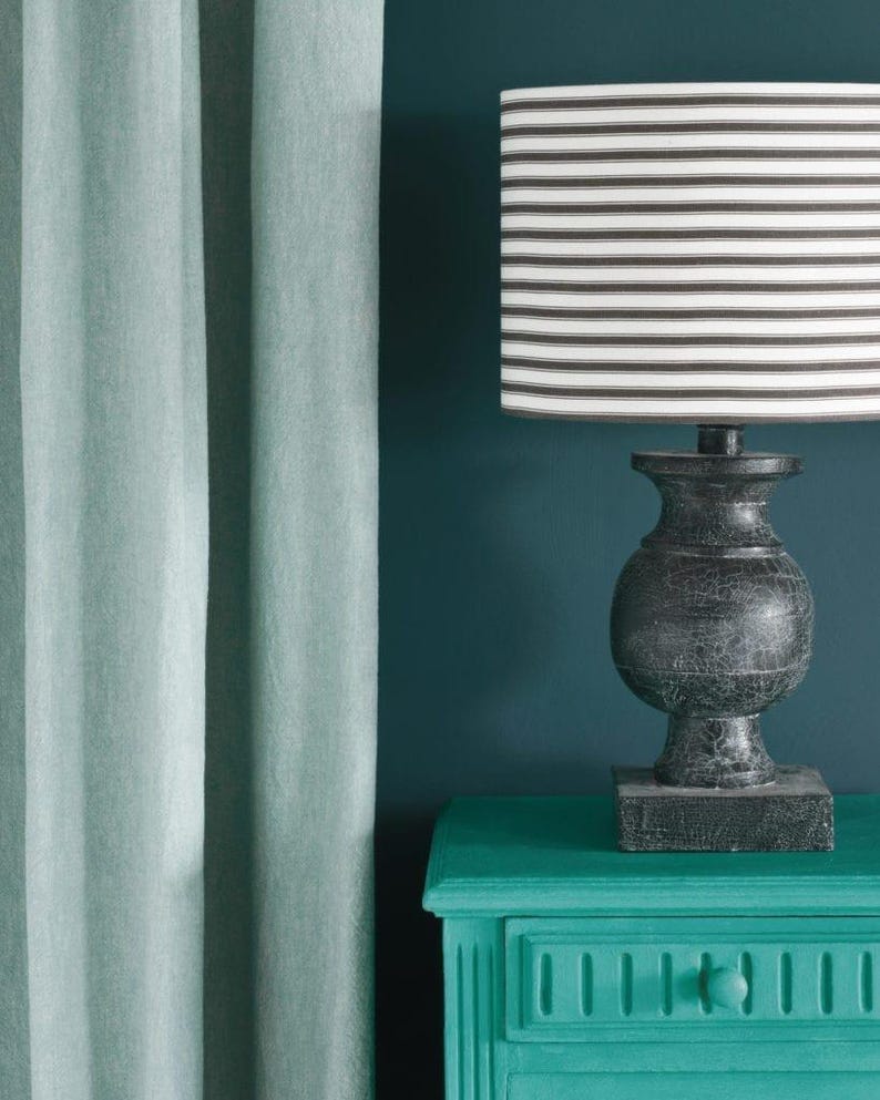 K&ouml;nnte beinhalten: Eine dekorative Lampe mit einem schwarz-wei&szlig; gestreiften Lampenschirm steht auf einem t&uuml;rkisfarbenen Nachttisch. Die Lampe hat einen dunklen, strukturierten Sockel. Weiche t&uuml;rkisfarbene Vorh&auml;nge sind links, der Hintergrund ist tief t&uuml;rkis.