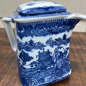May include: Blue and white ceramic teapot with a lid. The teapot features a detailed landscape design with buildings, mountains, and clouds. The handle and spout are white. The lid has a small knob.