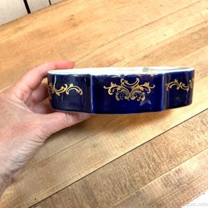 May include: A navy blue porcelain dish with a white interior and gold filigree designs. The dish has a unique, multi-faceted shape. The gold detailing adds a touch of elegance to this decorative piece.