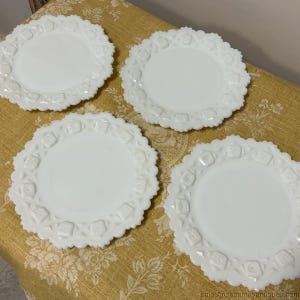 May include: Four white, vintage-style plates with a scalloped edge and embossed floral design. The plates are arranged on a yellow floral tablecloth, showcasing their delicate details and classic aesthetic. Each plate has a diameter of approximately 8 inches.