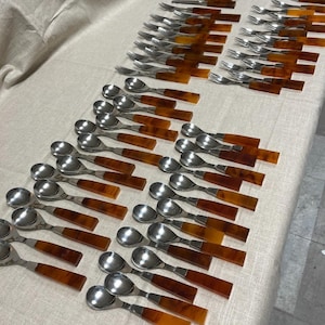 May include: An assortment of vintage silverware, including spoons and forks, arranged on a light-colored tablecloth. The silverware features stainless steel heads and handles with a tortoiseshell pattern. The arrangement suggests a formal dining setting.