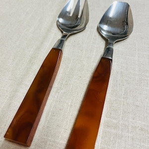 May include: A serving fork and spoon set with stainless steel heads and brown, rectangular handles. The fork has two tines. The spoon has a rounded bowl. The handles have a marbled appearance.
