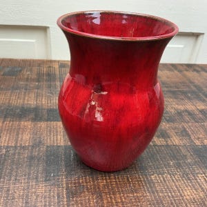 May include: A ceramic vase with a glossy, vibrant red glaze featuring subtle darker red and black undertones. The vessel has a wide, flared rim that tapers into a rounded, bulbous body, showcasing a smooth, reflective surface throughout the piece.