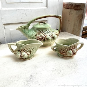 May include: A vintage ceramic tea set in shades of green and brown. The set includes a teapot with a curved handle, a creamer, and a small cup. The pieces feature a textured design with a botanical theme.