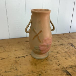 May include: A peach-coloured ceramic vase with two handles and a floral design. The vase features a pink flower and a brown 'X' design. The vase is approximately 20 cm tall.